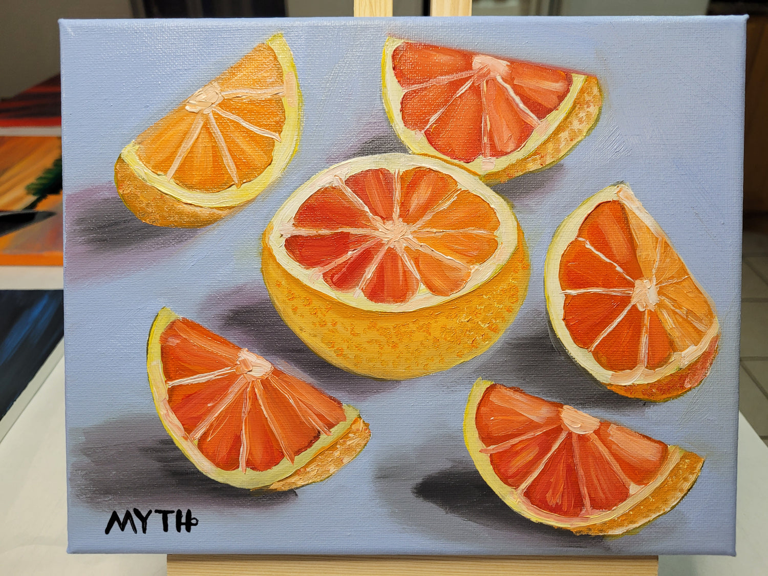 Oil on canvas painting of "Grapefruit" by artist MYTH. Grapefruit cut into 5 wedges neatly scattered around a half grapefruit that is face up in the center of the painting. The background is a cool blue and the grapefruits are shaded and shadowed appear as if the light is coming from the right side of the area. 